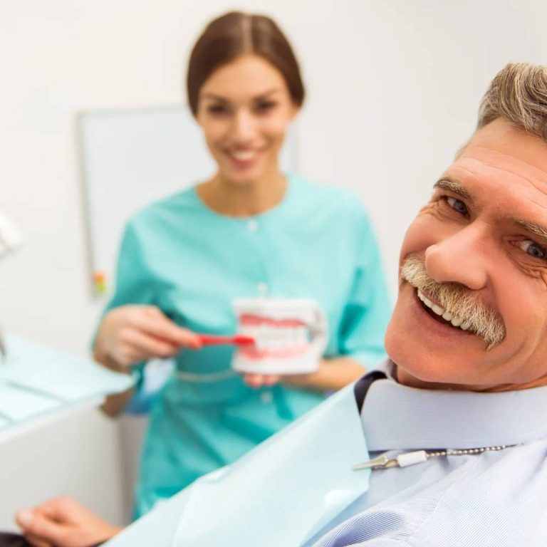 Middle-aged man smiling with female dental professional in turquoise scrubs holding tooth model in bright dental clinic
